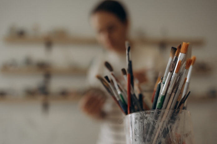 Artist supplies at wooden table. Paint brush in clay jug and art painter tools on wood desk background texture. Paintbrush for painting in artist studio workplace