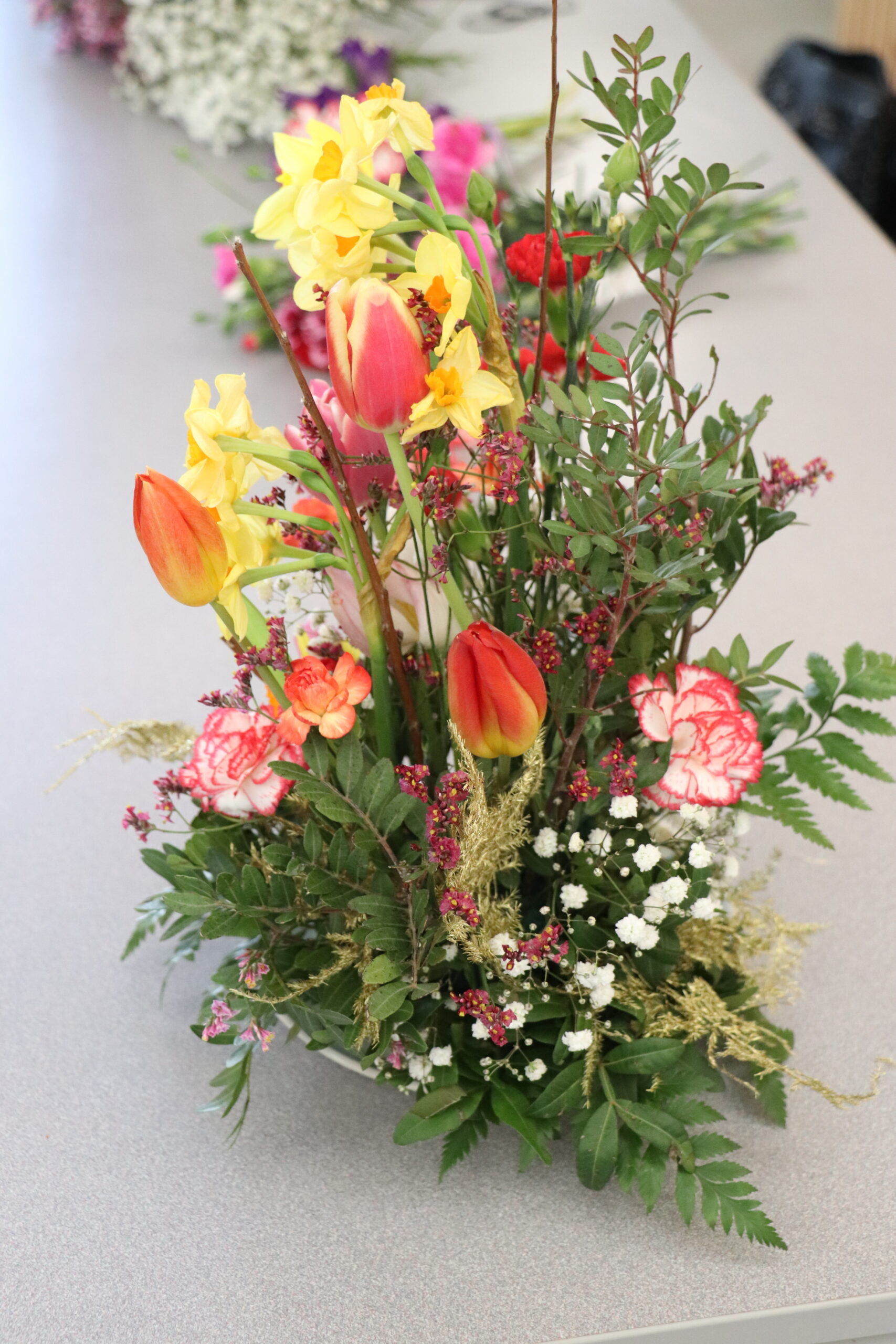 Composition florale printanière avec des tulipes, jonquilles et œillets sur fond de fougères.